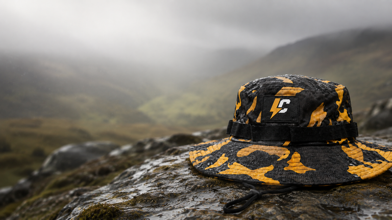 A camouflage bucket hat in black and yellow sits on a wet rock with a misty mountain landscape in the background. A small black patch on the side of the hat features a white stylized 'C' logo.